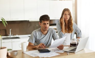 Concentrated young unshaven man dressed in t-shirt holding document, using calculator while paying taxes online on laptop pc, his frustrated wife standing behind him and having phone conversation