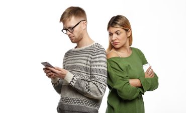 Studio shot of suspicious distrustful young blonde female in green top standing next to her bearded boyfriend, looking over his shoulder, spying over him while he is typing message using mobile phone